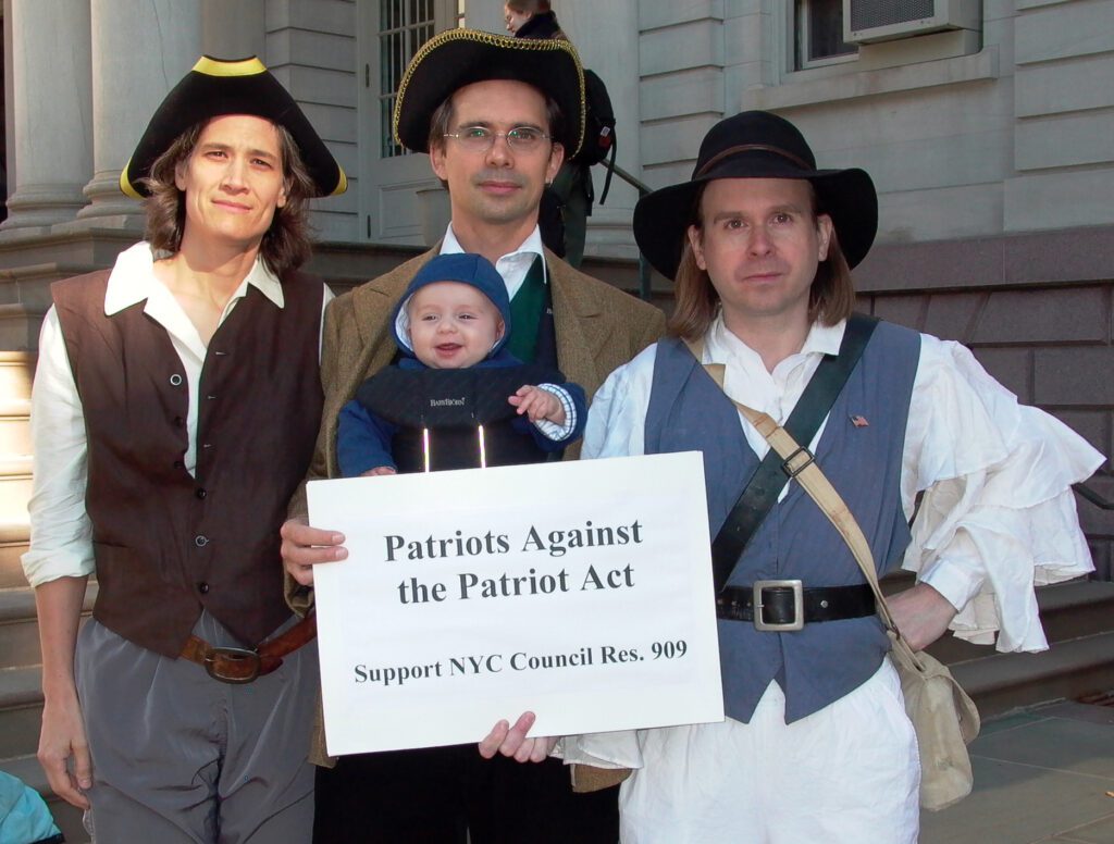 Demonstrators dressed as 18th Century American Revolutionaries demonstrate in front of City Hall in 2003. They favored a resolution before the New York City Council supporting restoration of civil liberties curtailed by the government since Sept. 11, 2001. Despite the City Council passing the resolution in 2004--and more than 250 other cities and three states, provisions of the Patriot Act remain in effect in 2022. Objections are still relevant. See https://www.aclu.org/other/surveillance-under-usapatriot-act. Left to right: Kelly Moore, Stephen Duncombe, Sydney Railla-Duncombe (aged 4 months), Bob Lesko. Photo by Pat Arnow